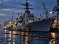 Photorealistic illustration of a U.S. warship undergoing shipyard maintenance at dusk, with a thin red harbour boom across the foreground.